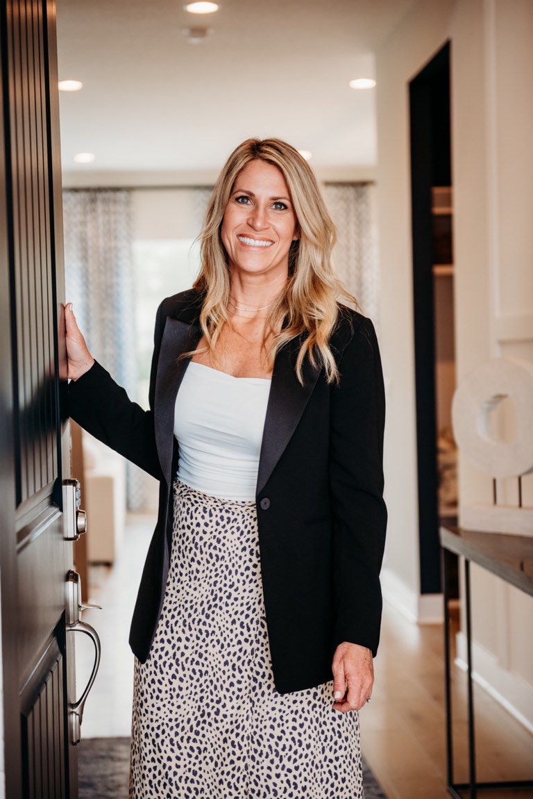 Becky Kampmeyer standing in the entry hall of a finished home with one hand on the open black front door, wearing a black blazer over a white top and a printed midi skirt, with a console table and light oak floors behind her