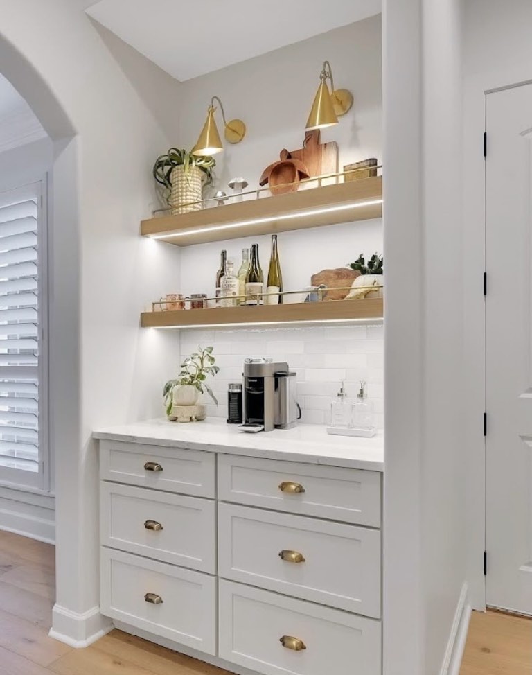 Built-in coffee bar with white shaker drawers, brass cup pulls, white quartz counter, subway tile and two white oak floating shelves with brass gallery rails
