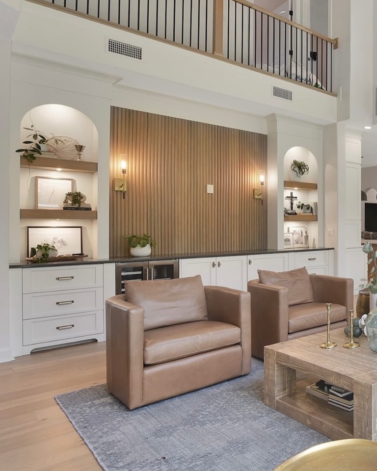 Two-story living room with a fluted white-oak feature wall between two lit arched niches, white cabinetry with a dark stone counter, brass plate sconces and two camel leather swivel chairs