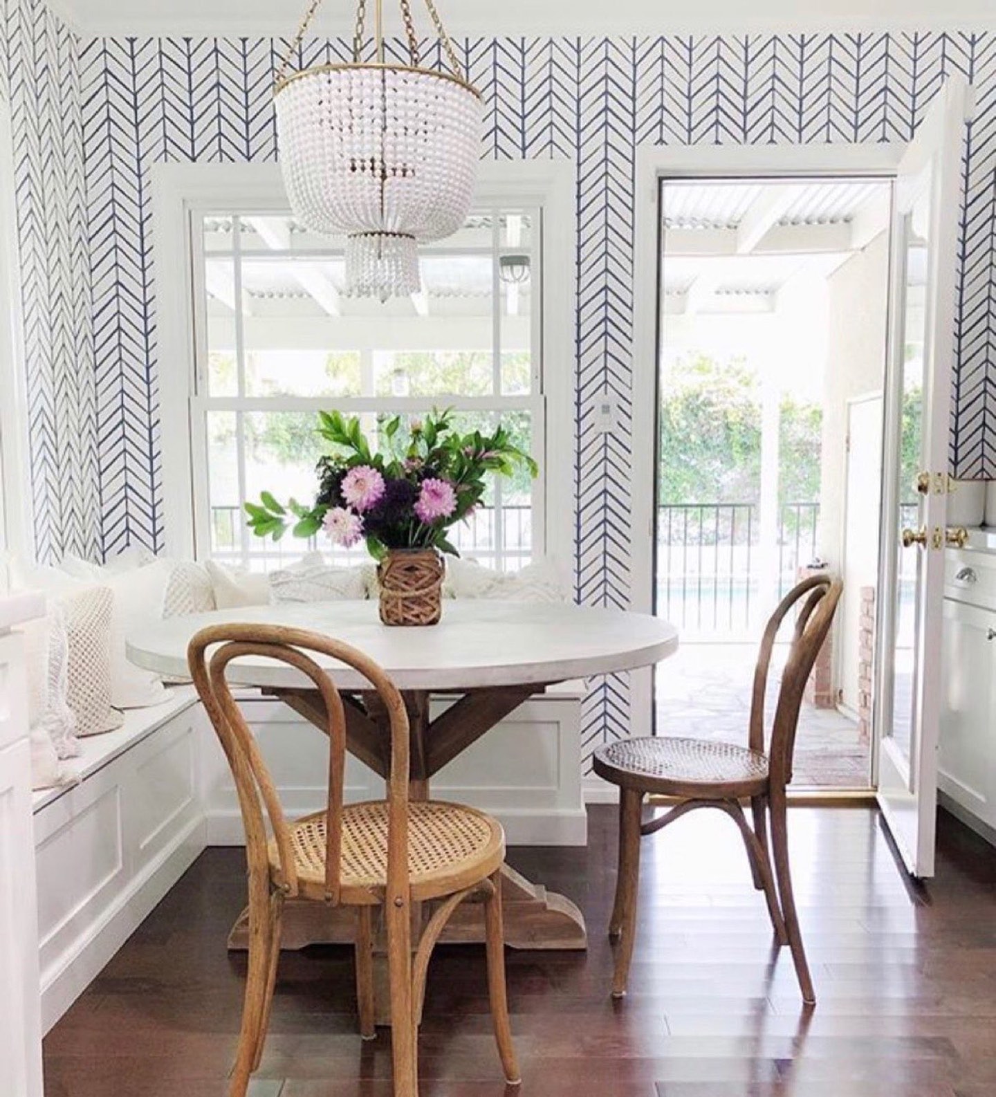 Breakfast nook papered in navy herringbone on white, with a beaded chandelier over a round trestle table and a built-in white banquette